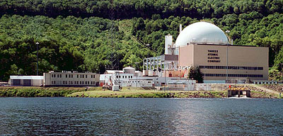 Photo of the plant during operation, showing admin/control building in front of the containment dome, and an array of other buildings on the waterfront, backed by green hills and a nascent power transmission line